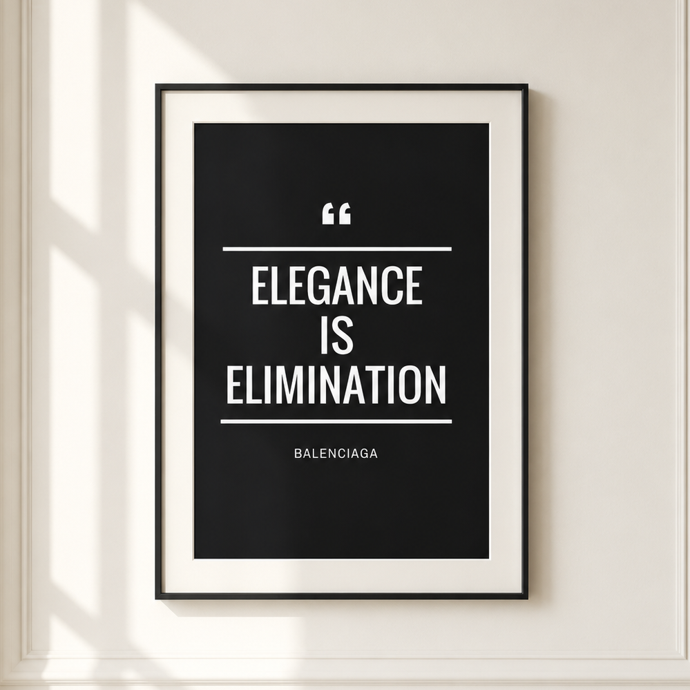 A framed minimalist quote print hanging on a cream-colored wall with decorative molding and soft window shadows. The print features a black background with white text reading 'ELEGANCE IS ELIMINATION' and the Balenciaga name at the bottom.