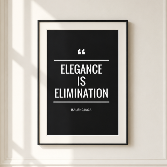 A framed minimalist quote print hanging on a cream-colored wall with decorative molding and soft window shadows. The print features a black background with white text reading 'ELEGANCE IS ELIMINATION' and the Balenciaga name at the bottom.