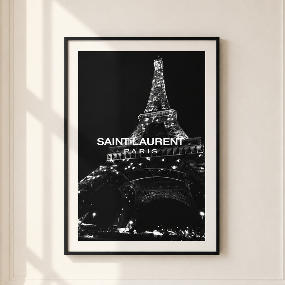 A framed black and white poster of the Eiffel Tower with 'SAINT LAURENT PARIS' text, displayed on a cream-colored wall with soft window shadows.