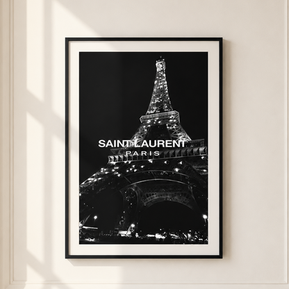 A framed black and white poster of the Eiffel Tower with 'SAINT LAURENT PARIS' text, displayed on a cream-colored wall with soft window shadows.