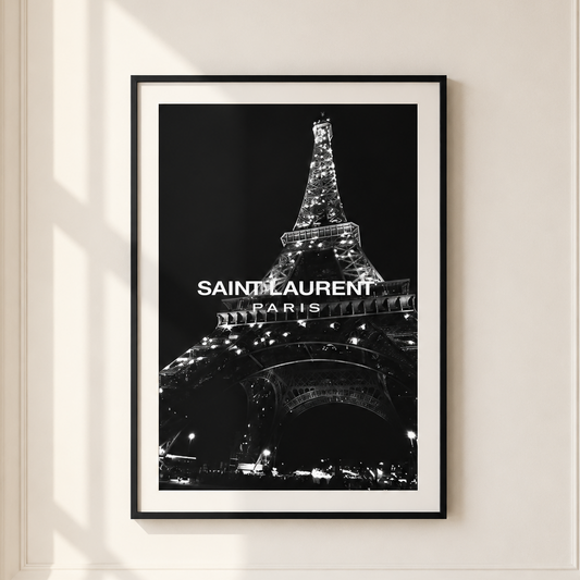 A framed black and white poster of the Eiffel Tower with 'SAINT LAURENT PARIS' text, displayed on a cream-colored wall with soft window shadows.