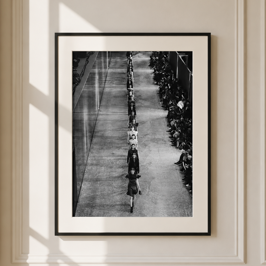 A framed black and white art print depicting a long line of people walking down a hallway, hung on a cream-colored paneled wall with soft window shadows.