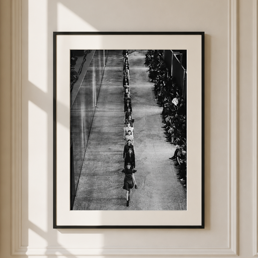 A framed black and white art print depicting a long line of people walking down a hallway, hung on a cream-colored paneled wall with soft window shadows.