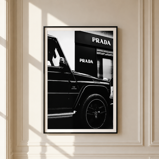 A framed black and white art print featuring a close-up of a black SUV in front of a Prada storefront, displayed on a cream-colored wall with decorative molding and soft window shadows.