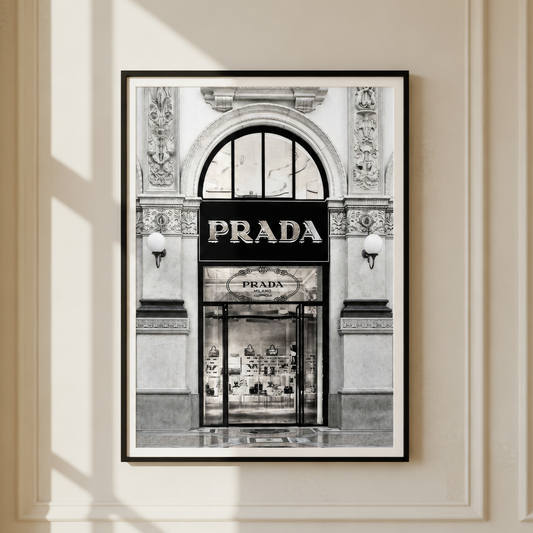 A framed black and white art print of a Prada storefront hanging on a cream-colored paneled wall with soft window shadows.