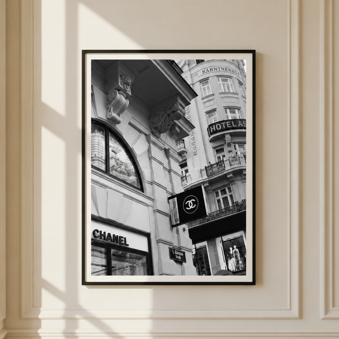 A framed black and white street art print featuring iconic Parisian architecture and storefronts, including a Chanel sign, hanging on a cream-colored paneled wall with soft window shadows.