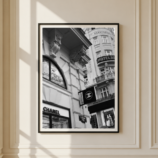 A framed black and white street art print featuring iconic Parisian architecture and storefronts, including a Chanel sign, hanging on a cream-colored paneled wall with soft window shadows.