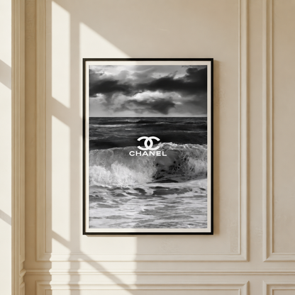 A framed black and white poster featuring a stormy ocean scene with the Chanel logo and text centered over the waves, hung on a cream paneled wall with soft window shadows.
