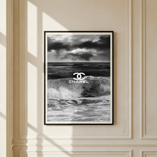 A framed black and white poster featuring a stormy ocean scene with the Chanel logo and text centered over the waves, hung on a cream paneled wall with soft window shadows.
