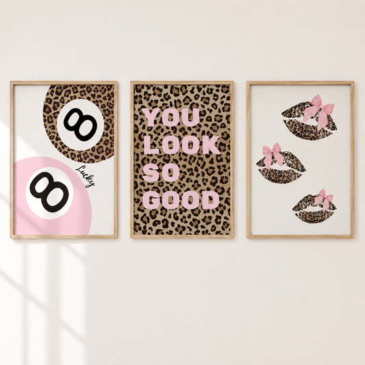 Three framed wall art prints with a leopard print theme hang on a light beige wall. The left print shows two black 8-balls on a pink and brown background with the word 'Lucky.