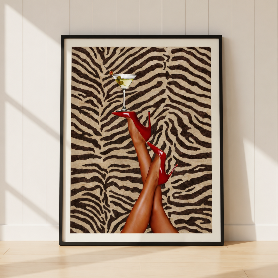 A framed art print leaning against a white paneled wall on a light wood floor. The print features a zebra-stripe pattern background with a pair of legs wearing red high heels and holding a martini glass.