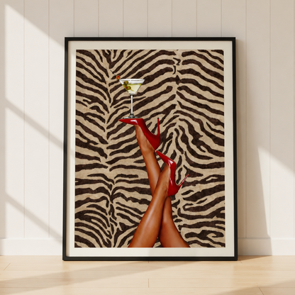 A framed art print leaning against a white paneled wall on a light wood floor. The print features a zebra-stripe pattern background with a pair of legs wearing red high heels and holding a martini glass.