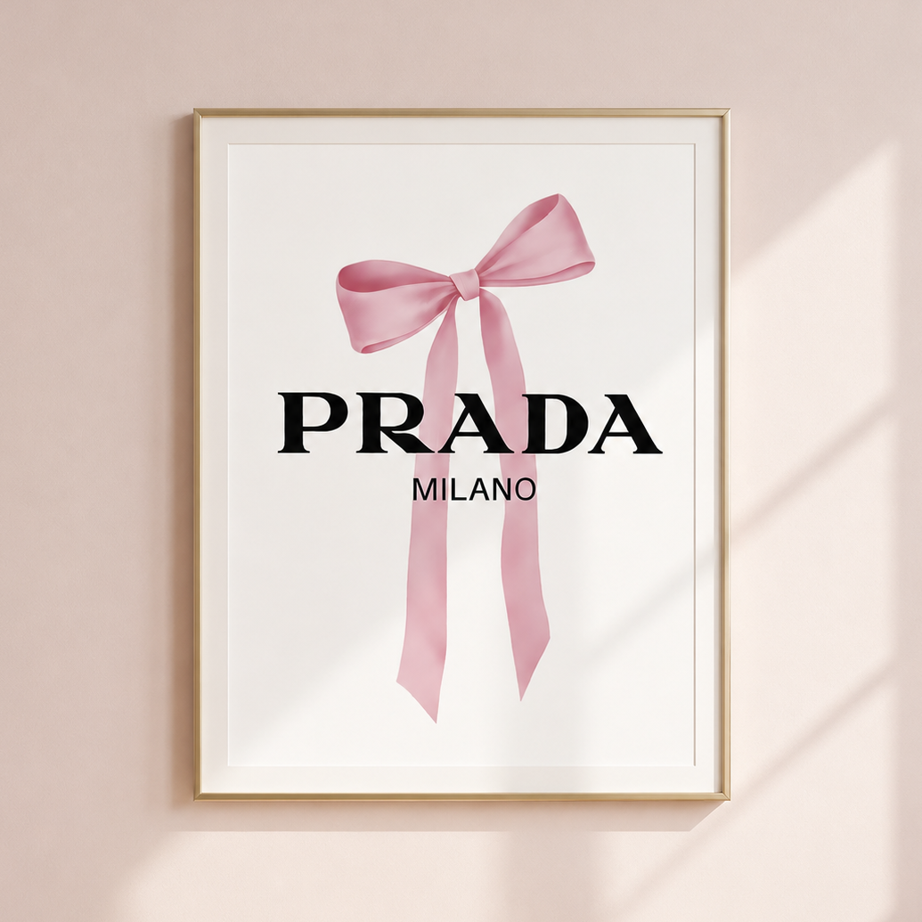 A framed wall art print featuring a pink ribbon bow and the text 'PRADA MILANO' on a white background, hung on a pale blush wall with soft window shadows.
