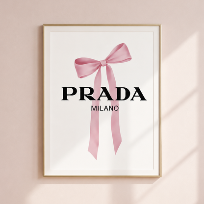 A framed wall art print featuring a pink ribbon bow and the text 'PRADA MILANO' on a white background, hung on a pale blush wall with soft window shadows.