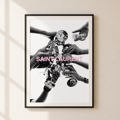 A framed black and white art print featuring multiple hands holding Saint Laurent perfume bottles, with the brand name printed in pink across the center, displayed on a beige wall with soft window shadow patterns.