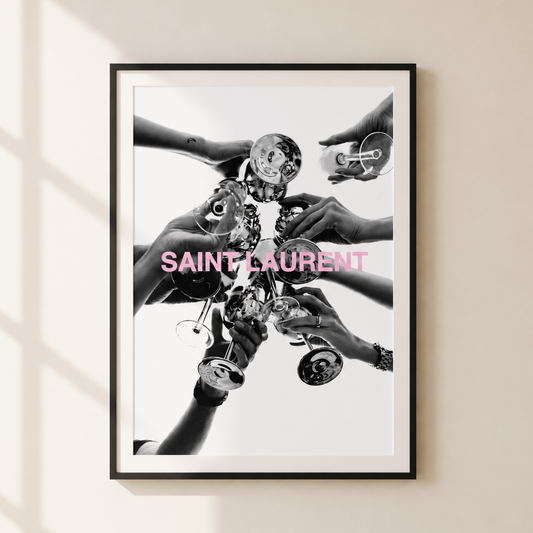 A framed black and white art print featuring multiple hands holding Saint Laurent perfume bottles, with the brand name printed in pink across the center, displayed on a beige wall with soft window shadow patterns.