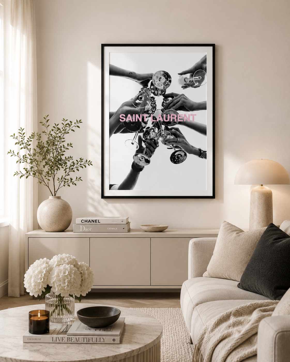 A framed black and white art print featuring multiple hands holding a camera, with pink 'SAINT-SAURENT' text across the center, displayed on a beige wall in a modern living room setting.