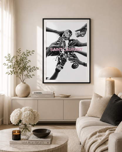 A framed black and white art print featuring multiple hands holding a camera, with pink 'SAINT-SAURENT' text across the center, displayed on a beige wall in a modern living room setting.