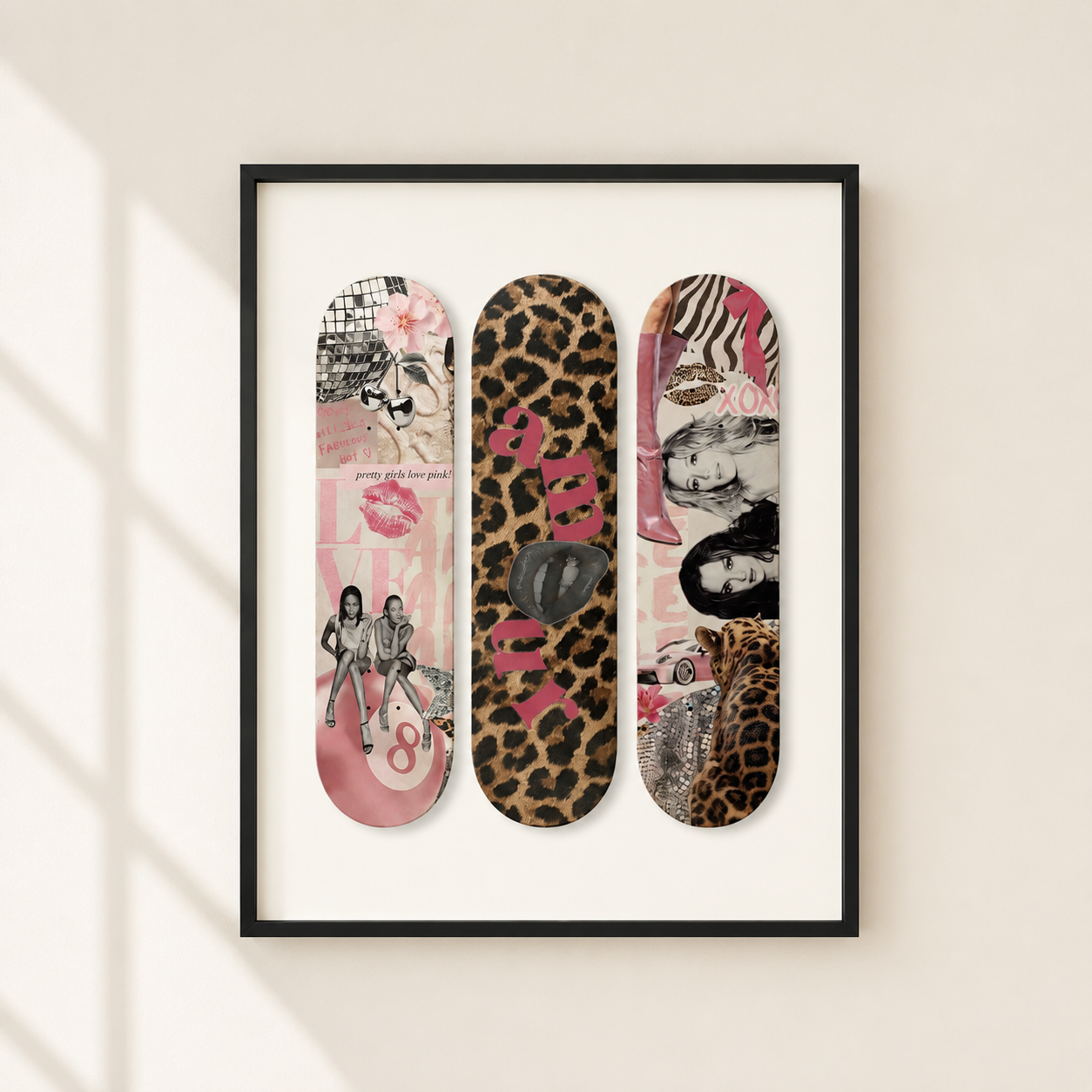 A framed wall art print featuring three vertical skateboard-deck style collage designs in pink, leopard print, and mixed-media tones, hung on a light beige wall with soft window-shadow lighting.