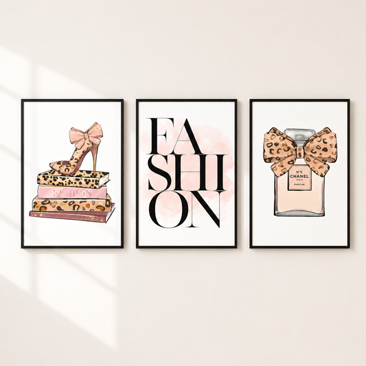 Three framed fashion-themed wall art prints displayed on a light beige wall. The left print shows a leopard-print high heel resting on stacked fashion books; the center print features the word 'FASHION' in large black letters with a soft pink watercolor splash; the right print shows a perfume bottle with a leopard-print bow and a small Chanel label.