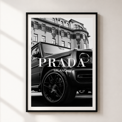 A framed black and white art print featuring a luxury car in front of an old building, with the text 'PRADA MILANO' overlaid across the center. The print is displayed on a light beige wall with soft window shadows.