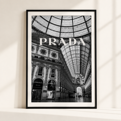 A black and white framed art print of the Galleria Vittorio Emanuele in Milan, featuring the word 'PRADA' in large letters across the center, displayed on a white shelf against a wall with soft window shadows.