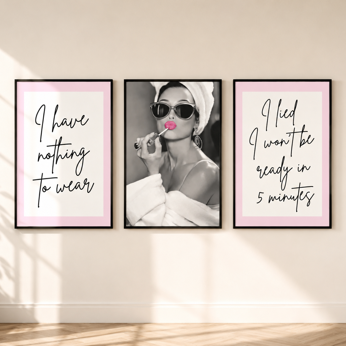 Three framed wall art prints displayed side by side on a beige wall above a wood floor. The left print has a pink border, Monochrome portrait of Audrey Hepburn with pink lipstick and black script text reading 'I have nothing to wear.