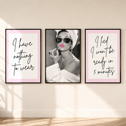 Three framed wall art prints displayed side by side on a beige wall above a wood floor. The left print has a pink border, Monochrome portrait of Audrey Hepburn with pink lipstick and black script text reading 'I have nothing to wear.