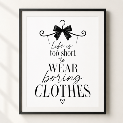 A framed typographic wall art print with the quote 'Life is too short to WEAR boring CLOTHES' in black text on a white background, with a black hanger and bow illustration at the top. The frame is black and mounted against a light neutral wall.