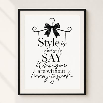 A framed typographic wall art print with a black hanger and bow illustration at the top and the quote, 'Style is a way to SAY Who you are without having to speak,' in black lettering on a white background. The frame is shown hanging against a light neutral wall.