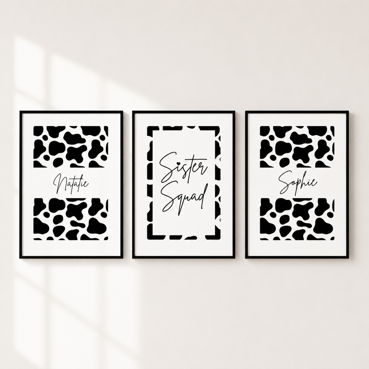 Three framed black and white cow print wall art prints hanging side by side on a light wall. The left and right prints feature black and white cow spots with the names 'Notica' and 'Sophie' in script, and the center print has a white background with a black border and the words 'Sister Squad' in script.