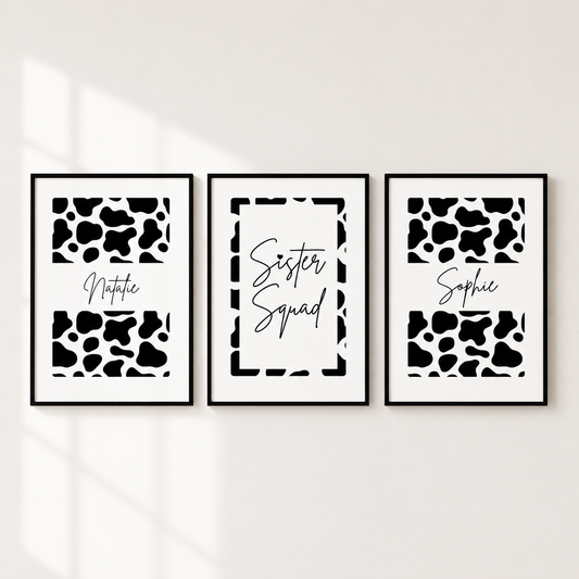 Three framed black and white cow print wall art prints hanging side by side on a light wall. The left and right prints feature black and white cow spots with the names 'Notica' and 'Sophie' in script, and the center print has a white background with a black border and the words 'Sister Squad' in script.