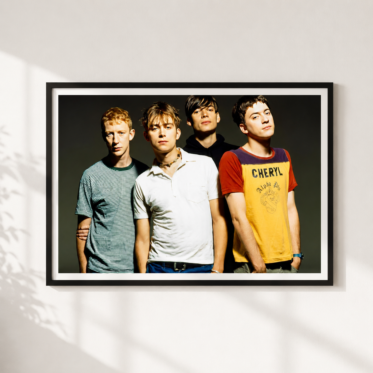 A framed black-and-white band portrait print hanging on a white wall, showing four young men in casual clothing against a dark background. The frame is black with a white mat, and the wall has soft window-shadow lighting.