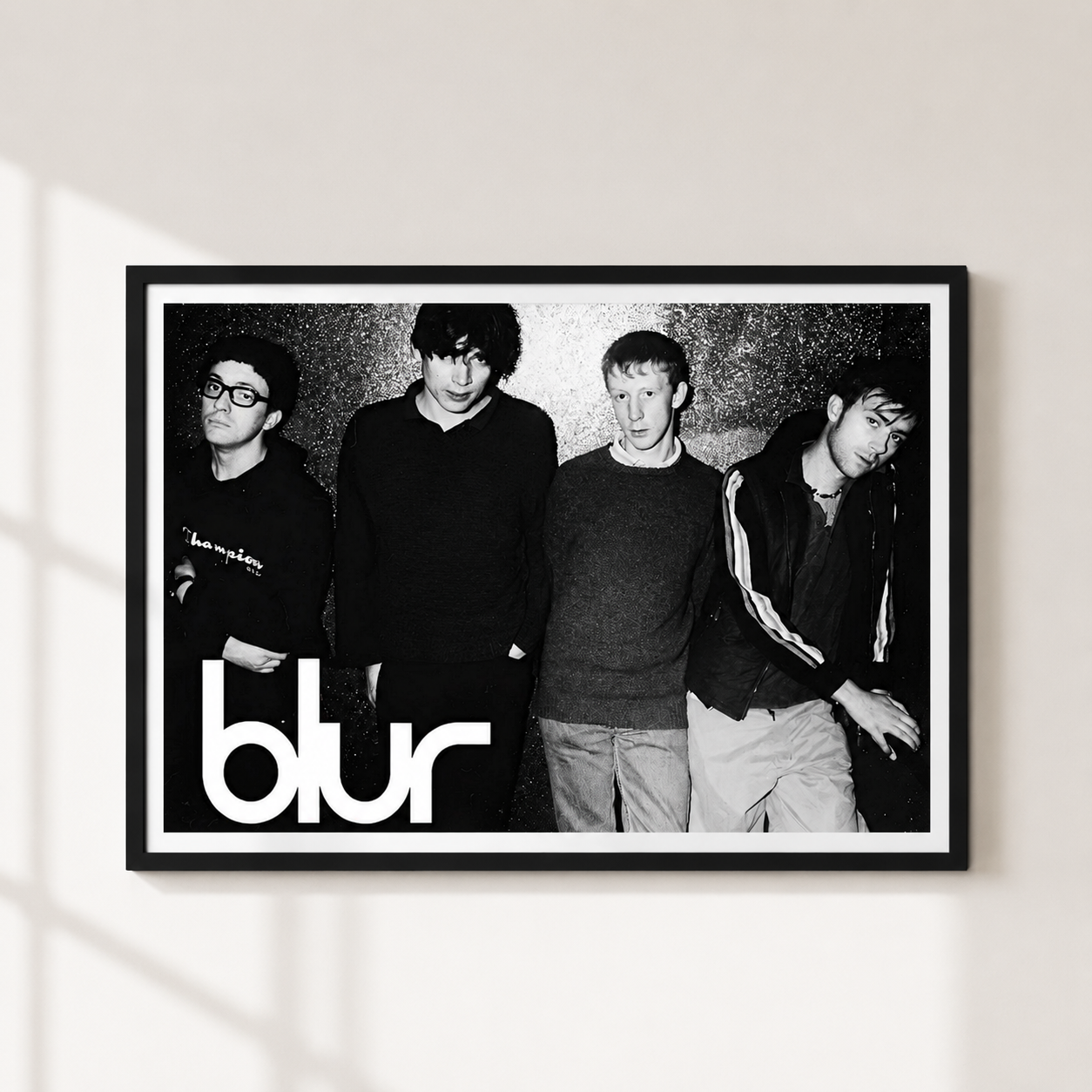 A framed black and white band poster of the music group 'Blur' hanging on a light beige wall. The poster features a black-and-white photo of the four band members with the band's name in large white lowercase letters at the bottom.