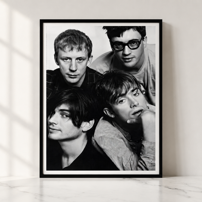 A framed black and white portrait print of the band Blur leaning against a light wall on a marble surface, with soft window shadows cast across the scene.