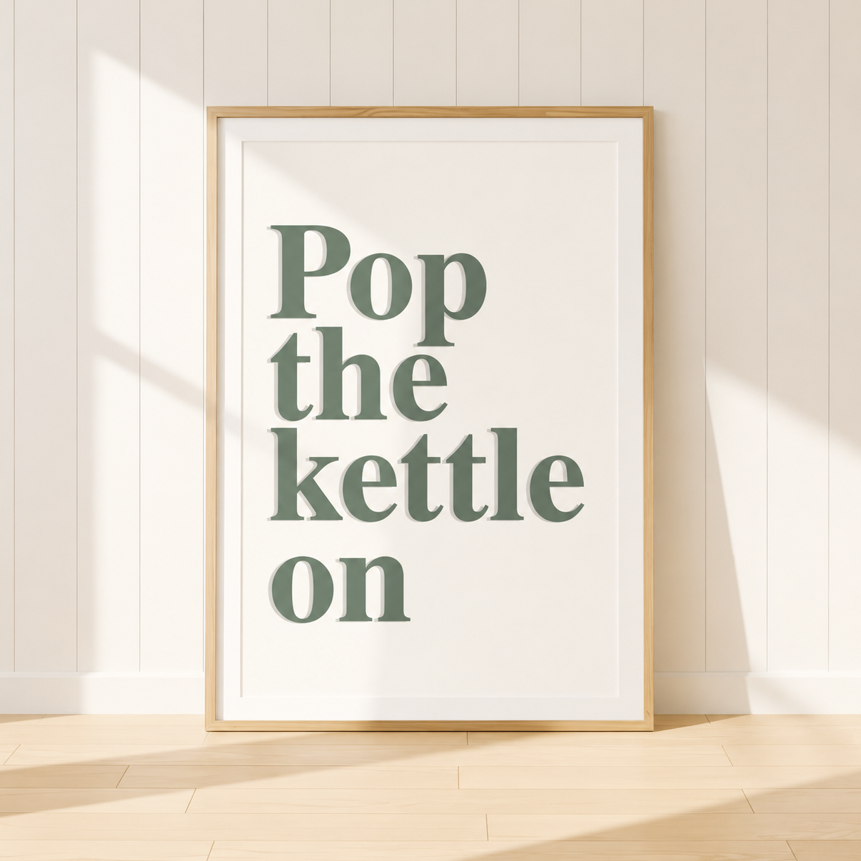 Framed "Pop The Kettle On" typography wall art in sage green with a light oak frame and warm ivory mount, styled in a bright modern farmhouse kitchen with natural wood accents and neutral décor.