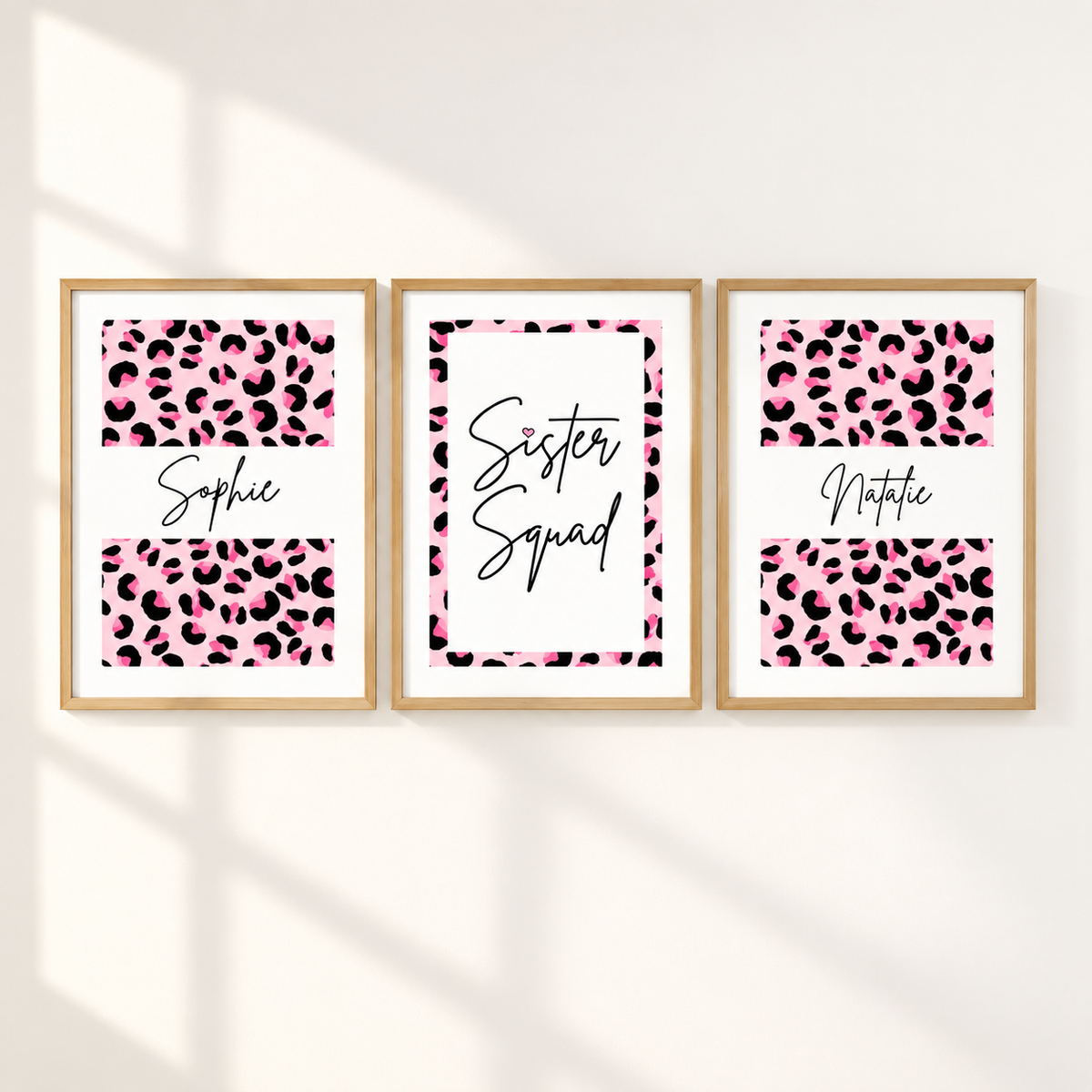 Three framed personalized prints with pink leopard print and black text hang on a white wall. The left print says 'Sophie', the center says 'Sister Squad', and the right says 'Noticia'.
