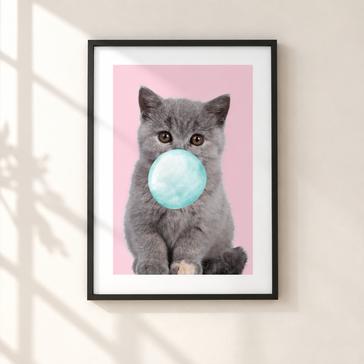 A framed art print featuring a gray kitten blowing a light blue bubblegum bubble against a pink background, displayed on a beige wall with soft window shadow overlays.