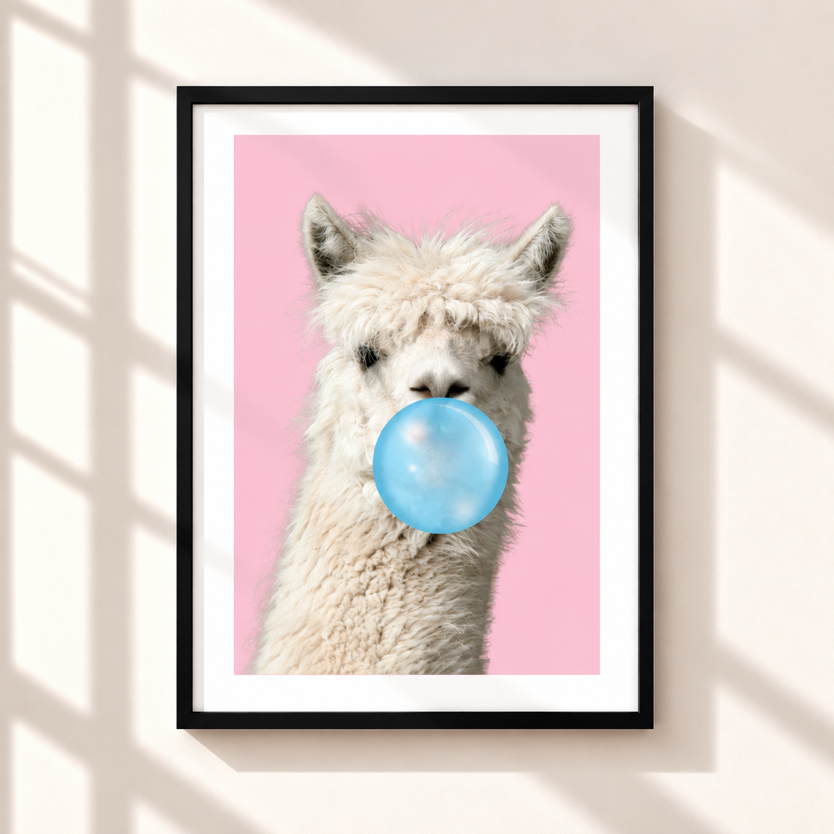 A framed art print featuring a close-up of a fluffy white alpaca or llama blowing a light blue bubblegum bubble against a solid pink background, displayed on a warm beige wall with soft window shadow overlays.