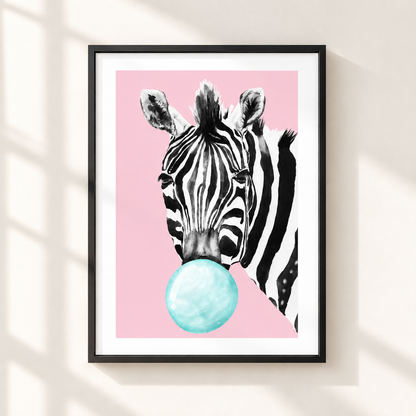 Framed wall art featuring a black-and-white zebra head blowing a light blue bubblegum bubble against a pink background, shown hanging on a beige wall with soft window-shadow lighting.