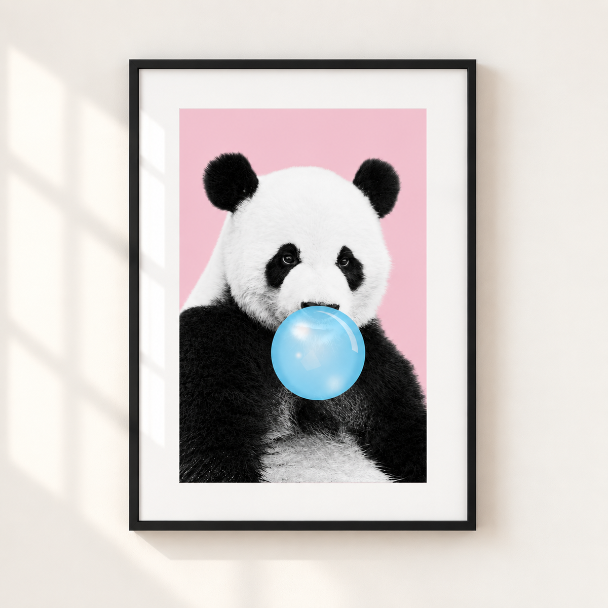 A framed art print featuring a black and white panda holding a light blue bubblegum bubble against a solid pink background, mounted on a light beige wall with soft window shadow overlays.