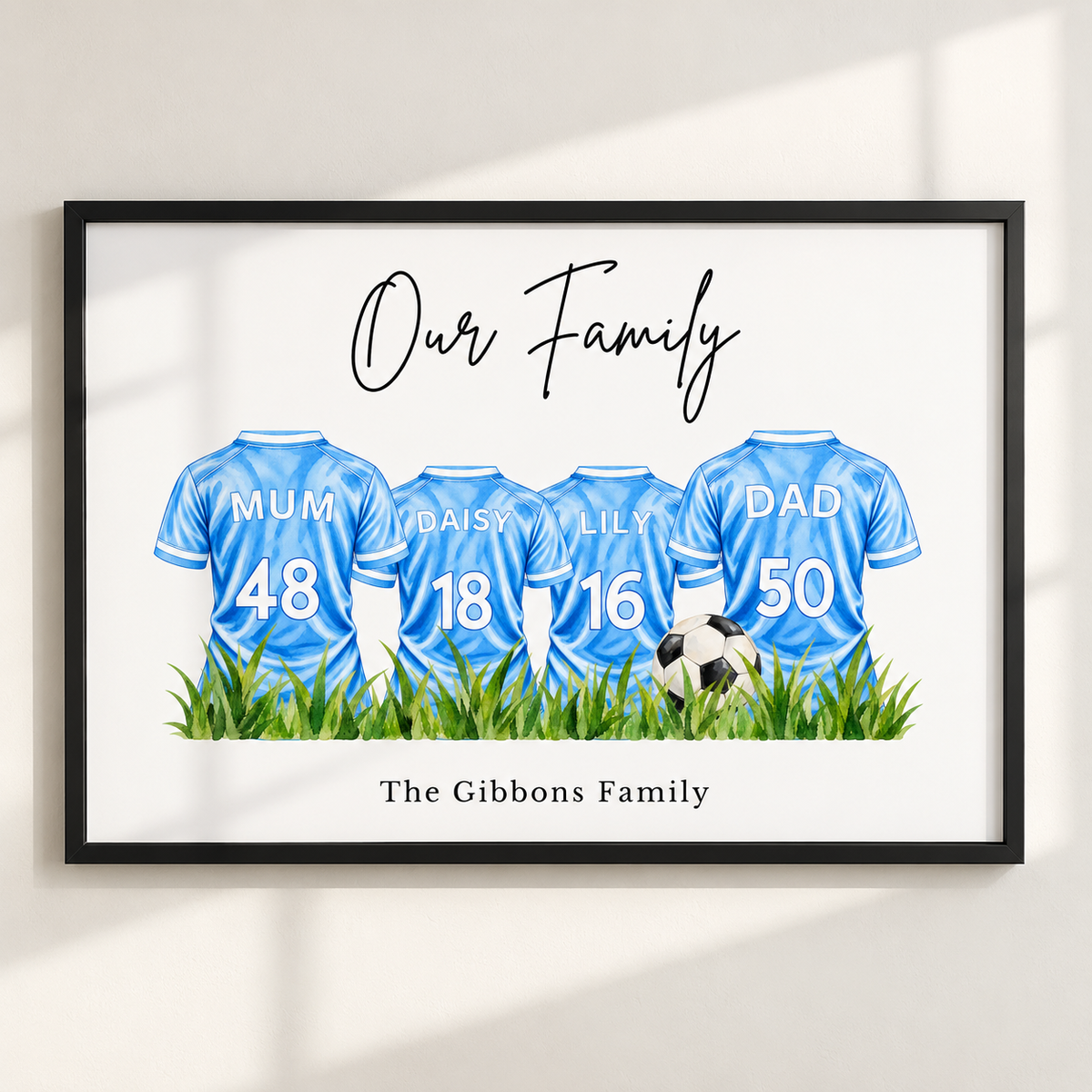 A framed personalized family art print featuring four light blue soccer jerseys with names and numbers for Mum, Daisy, Lily, and Dad, plus a soccer ball and grass. The print reads 'Our Family' at the top and 'The Gibbons Family' at the bottom, shown hanging on a light wall with soft window shadows.