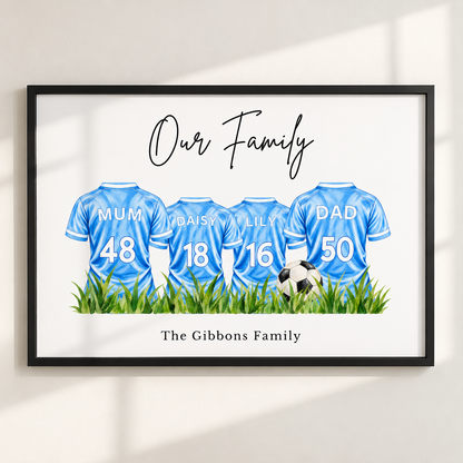 A framed personalized family art print featuring four light blue soccer jerseys with names and numbers for Mum, Daisy, Lily, and Dad, plus a soccer ball and grass. The print reads 'Our Family' at the top and 'The Gibbons Family' at the bottom, shown hanging on a light wall with soft window shadows.