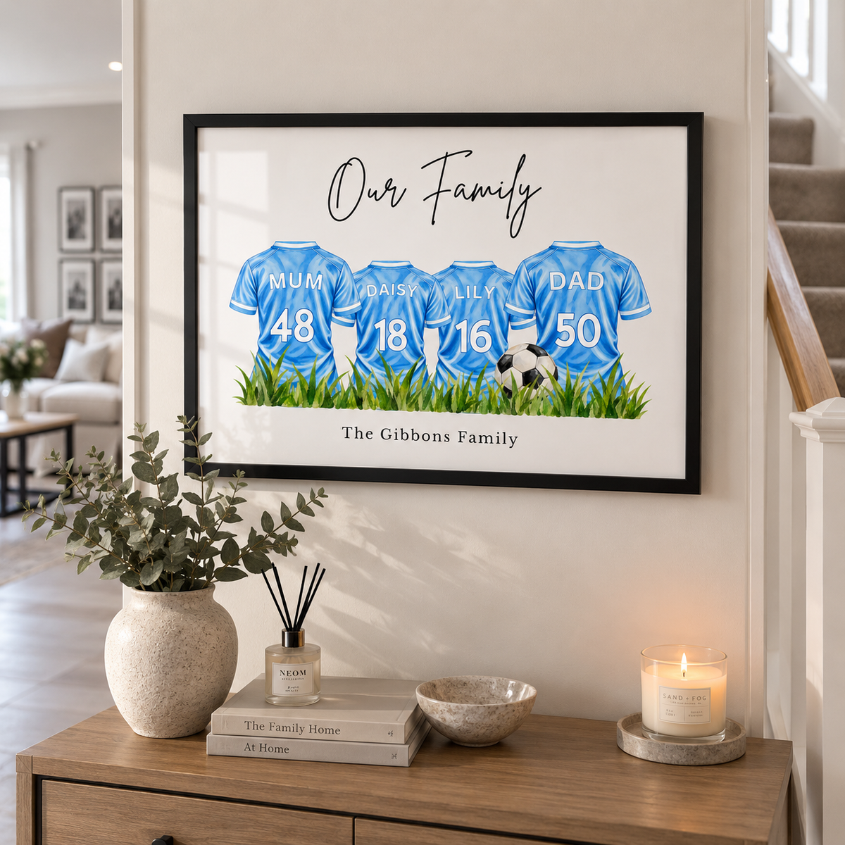 A framed custom family print hanging on a wall in a styled home interior. The print shows four blue football jerseys with the names Mum, Daisa, Ail, and Dad, plus numbers 48, 18, 16, and 50, above green grass and the text 'Our Family' and 'The Gibbons Family'.