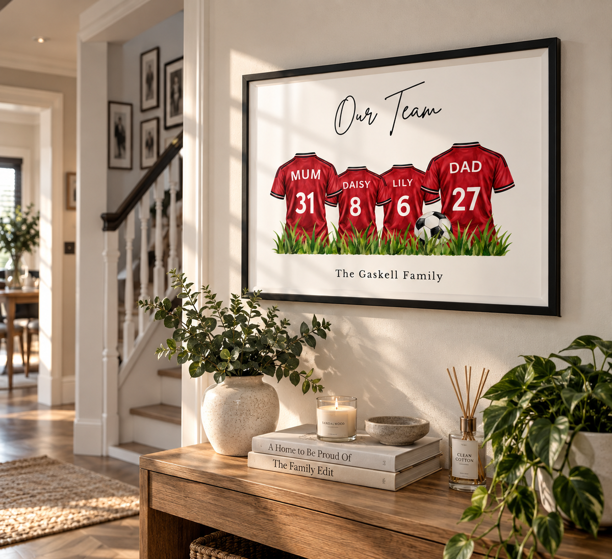 A framed custom family sports print hanging on a wall in a bright home interior. The artwork shows three red football jerseys labeled Mum, Raissy, Lily, and Dad with numbers 31, 8, 6, and 27, plus the text 'Our Team' and 'The Gaskell Family'.