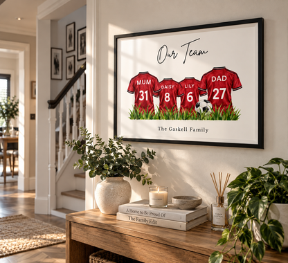 A framed custom family sports print hanging on a wall in a bright home interior. The artwork shows three red football jerseys labeled Mum, Raissy, Lily, and Dad with numbers 31, 8, 6, and 27, plus the text 'Our Team' and 'The Gaskell Family'.