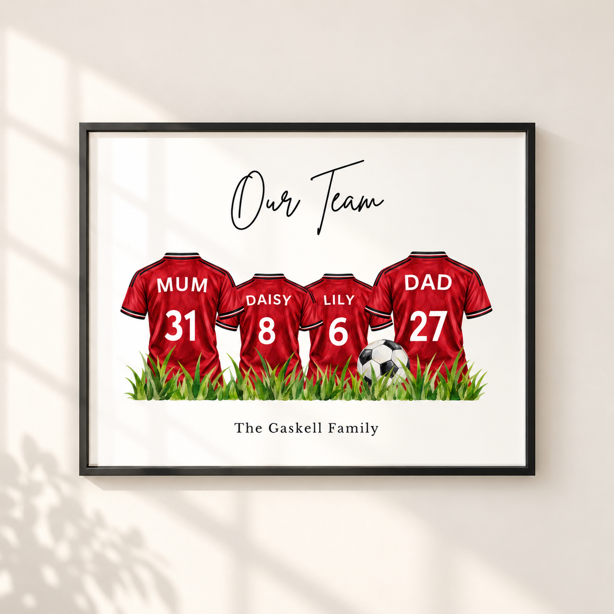 A framed personalized family football jersey wall art print hanging on a light beige wall. The print features four red soccer jerseys with the names 'MUM', 'DAISY', 'LILY', and 'DAD' and numbers 31, 8, 6, and 27 on the backs, standing on green grass with a small soccer ball.
