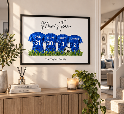 A framed custom family print hanging on a wall in a bright living room. The print shows four blue soccer jerseys with the names DAD, MUM, JOEY, and AURTHUR behind them, under the text 'Mum's Team' and above 'The Taylor Family'.