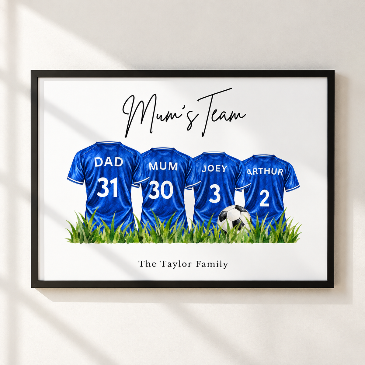 A framed personalized wall art print showing four blue soccer jerseys with names and numbers: Dad 31, Mum 30, Joey 3, and Arthur 2, above green grass and a soccer ball. The print includes the script text 'Mum's Team' and the family name 'The Taylor Family' at the bottom, displayed on a light wall with soft window shadows.