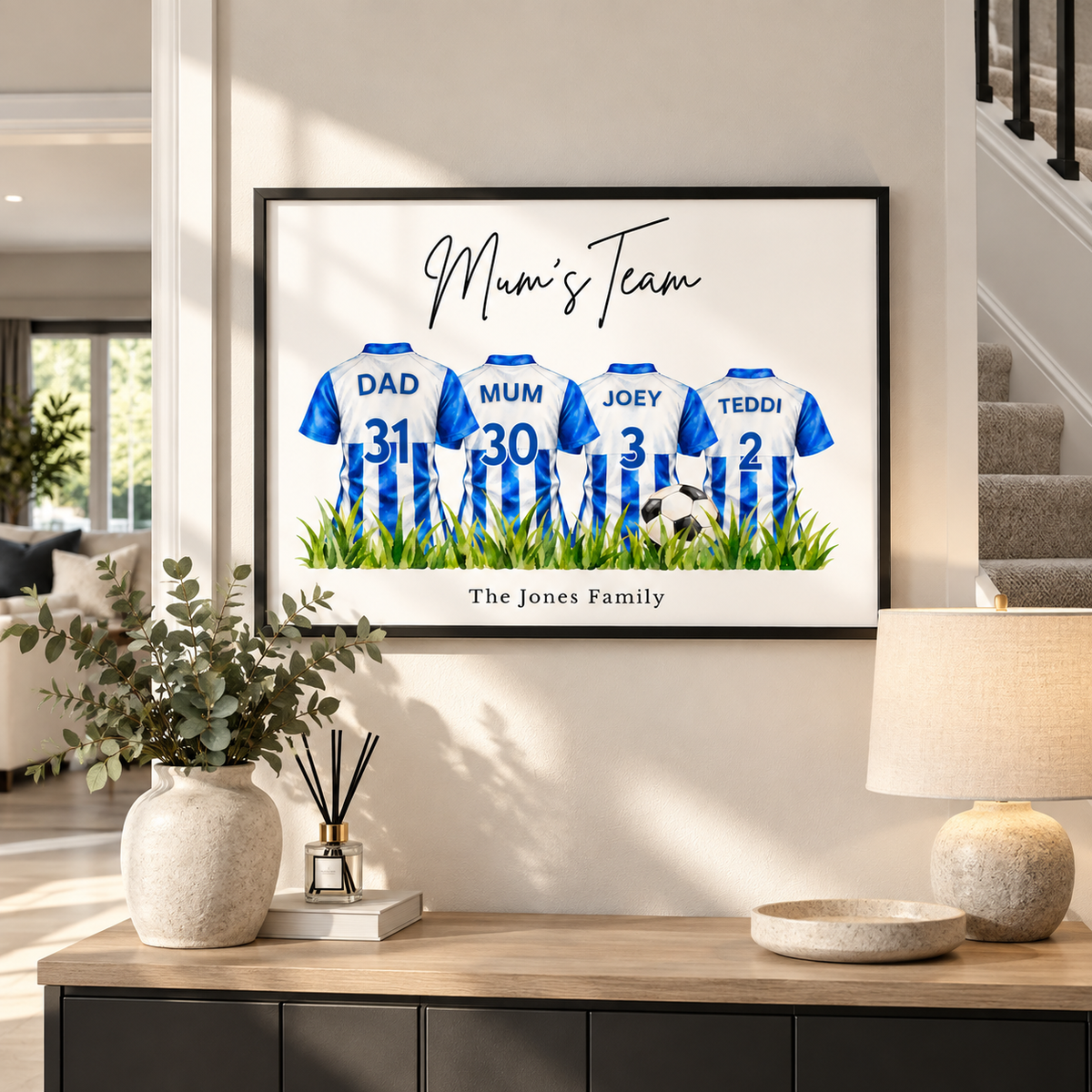 A framed custom family soccer jersey wall art print hanging on a wall in a modern living room. The print features blue and white striped soccer jerseys for Dad, Mum, Joey, and Teddi with the names 'The Jones Family' printed at the bottom.